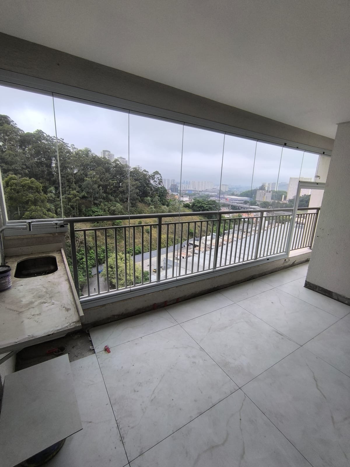 Sacada envidraçada com perfis e guarda-corpo brancos, pia à esquerda e vista para árvores e prédios